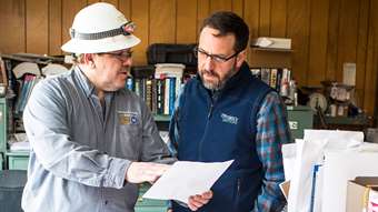 Two men, one in a hard hat and one with an Efficiency Vermont vest, talk while looking at a document