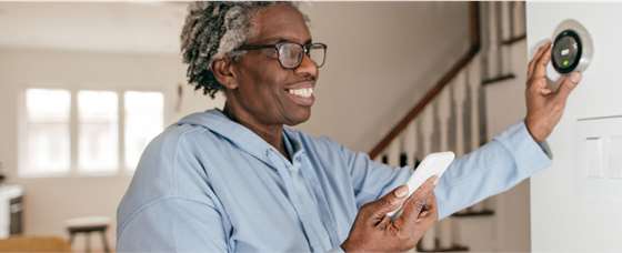 person of color with a smart phone adjusting a smart thermostat on the wall of their home