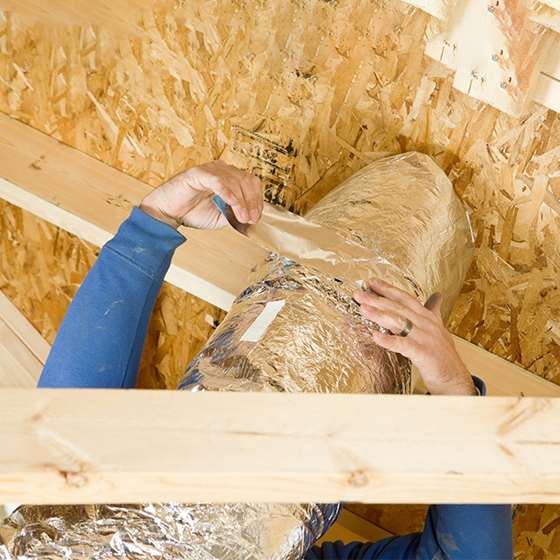 a pair of hands wrapping foil insulating tape around a duct in an attic