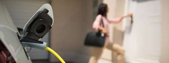 A plug going into a vehicle in the foreground, a white woman opens a door in the background