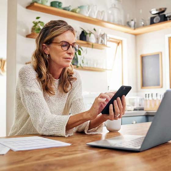blonde woman in glasses looking at a smart phone near a laptop
