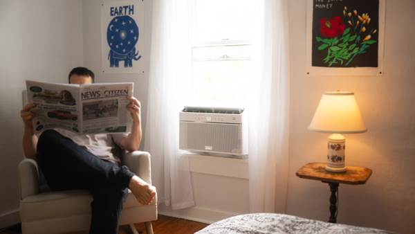 person sitting in a room reading a paper while an air conditioner sits in the window nearby