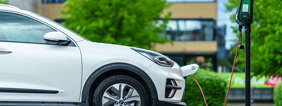white car with a electric plug in front bumper parked in front of an EV charger, green trees and buildings in the background