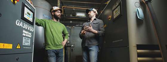 Two men stand near brewery equipment talking