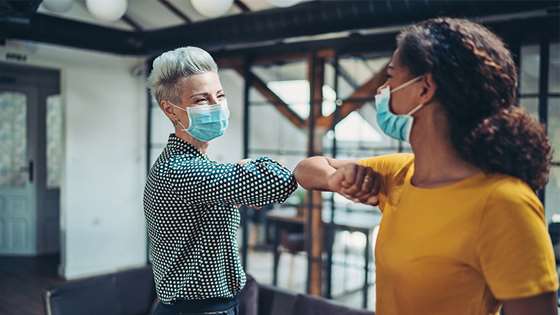 two women with masks for covid safety on touching elbows in a casual office setting