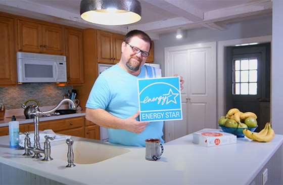 man stands in a brightly lit kitchen holding a sign with the ENERGY STAR label