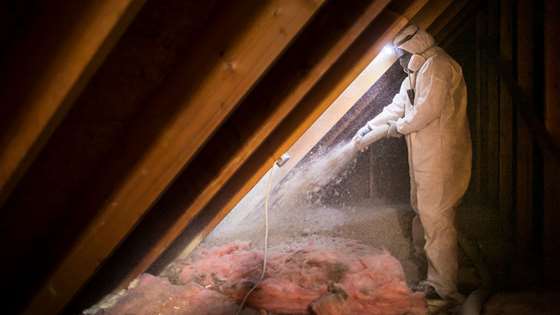 person in protective gear with hose and insulation blowing around an attic