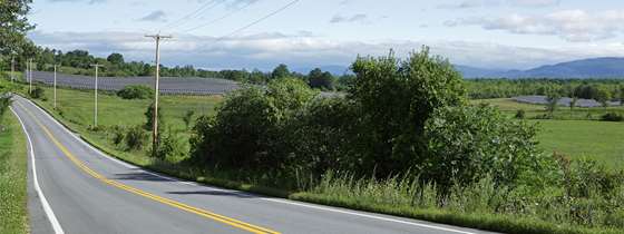 a rural two lane paved road with power lines and solar panels in view, rolling Vermont hills and green meadows are in background 