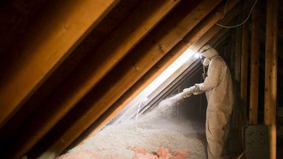 A contractor wearing white protective equipment blows cellulose insulation into a dark attic space