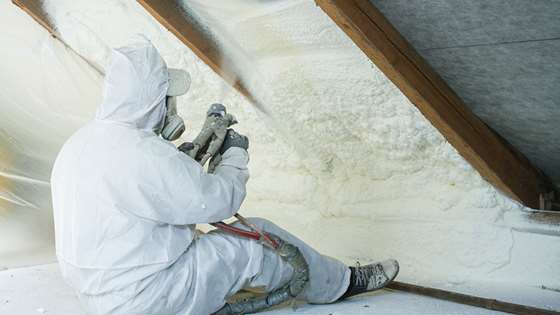 a contractor in full-body protective equipment sits in an attic applying spray foam insulation