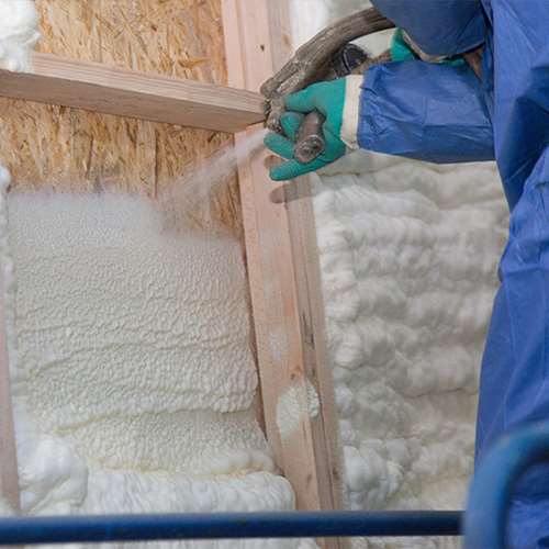 a contractor wearing green gloves and a blue protective suit applies spray foam to a wooden wall