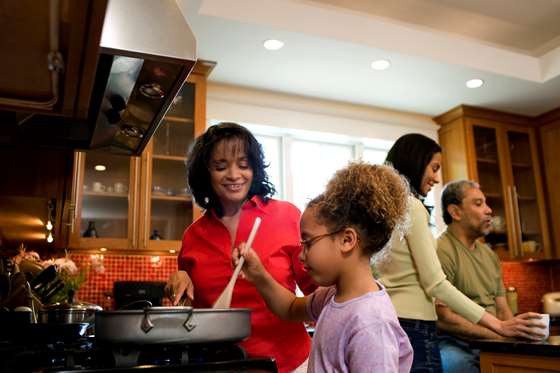 An older woman of color stands near a young black girl stirring a pot on a stove. In the background, a woman and a man have a conversation.