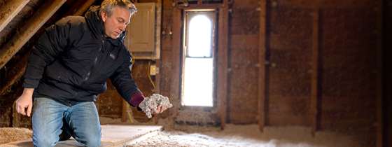 middle age man in jeans and black jacket kneeling in an attic to inspect blown in insulation