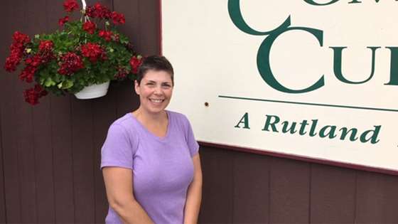 Rebekah Stephens, Executive Director Rutland Community Cupboard. She has short, dark hair and is wearing a light purple t-shirt. She is standing next to the Rutland Community Cupboard sign.