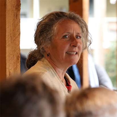 A woman addresses a crowd gathered under a timber-frame porch