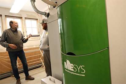 Two men talk in the background as a large new green and gray wood pellet boiler stands in the foreground.