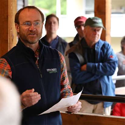 A bearded man in red flannel and a navy Efficiency Vermont vest points to a component of an indoor heating system.