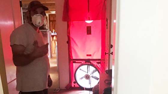 Jake Robichaud, wearing a mask, points at the blower door which is set up at Jill & Flynn's home