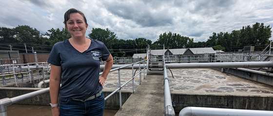 a woman standing on a concrete walkway at a waste water treatment facility, showing pipes and water in background