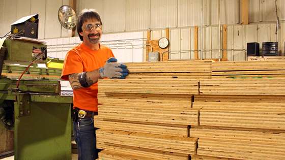 male employee in safety goggles and orange shirt on a manufacturing floor with plywood and machinery