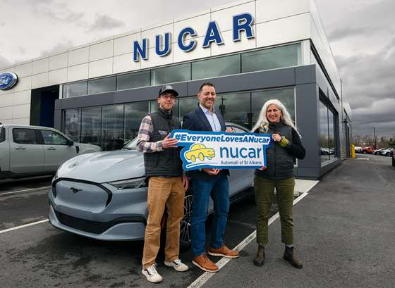 2 men and one woman stand in fron of a car dealership holdogn a sign reading Nucar