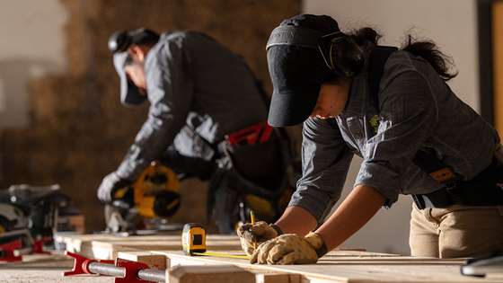 construction worker in the middle of a job site leaning over lumber with a measuring tape and pencil 