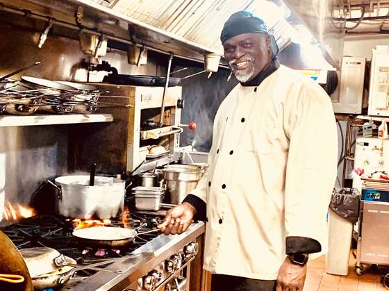 Frank Stone, head chef wearing a white chef's coat and a blue cap on his head smiles at the camera while standing in front of a stove. He's holding a frying pan over an open flame.