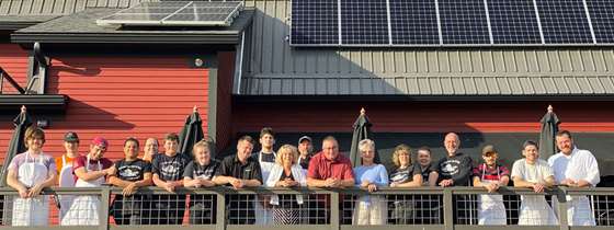 Photo looking up at a porch on the side of Publyk house. Chefs in white aprons, waitstaff in black t shirts, and the owners stand on the porch smiling down at the camera. Above them is the roof of the restaurant covered in solar panels.