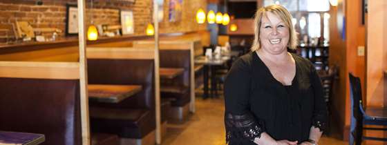 Photo of Kristin Murphy, co-owner of Twiggs, wearing a black shirt and with short blond hair and a big smile, standing in front of dining booths with softly lit hanging lamps over them