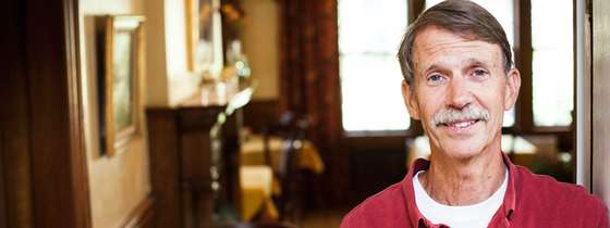 Dan, co-owner of Swift House, stands against the doorway to the inn's dining room where three small tables with white tablecloths are visible. Dan is smiling, wears a red collared shirt, and has dark gray-brown hair and a gray mustache.