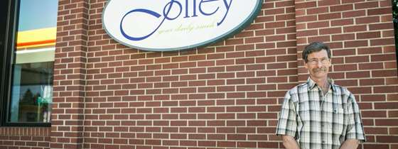 Photo of owner of Jolley gas station wearing a plaid t shirt and glasses standing in front of brick wall with Jolley sign on front