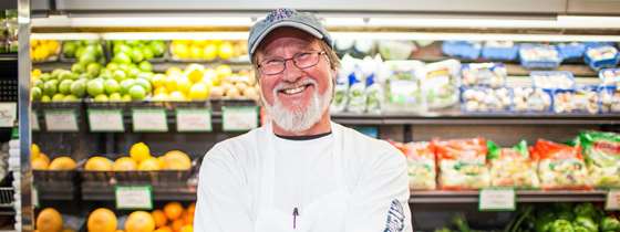 Photo of a man with glasses and a white beard smiling in front of refrigerator shelves containing grapefruit, lemons, limes, oranges, mushrooms, and green vegetables