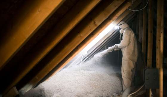 A person in safety gear sprays insulation in the attic of a home