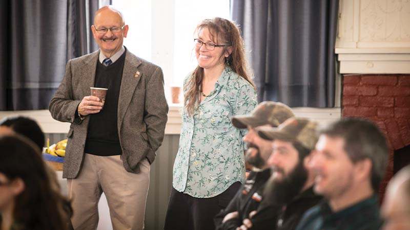 a man smiling holding a paper coffee cup stands next to a smiling woman in a room of seated people