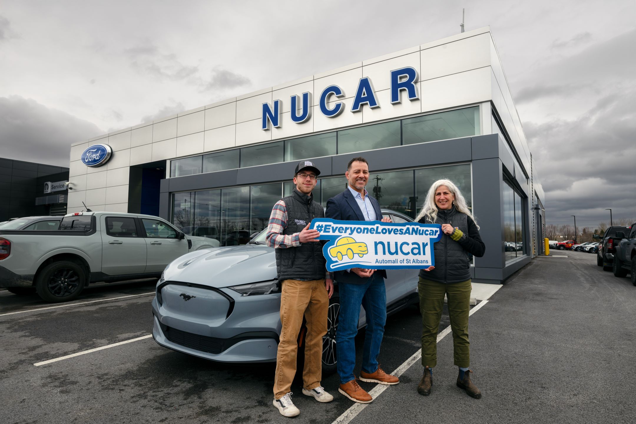 Efficiency Vermont's Alex Frei, left, and Nucar Automall General Manager Ryan Ortiz, middle, hold a Nucar logo with Efficiency Vermont's Nicole Bourassa, right. Nucar Automall in St Albans installed 15 new EV chargers through Efficiency Vermont's EV dealer program to support the selling and servicing of EVs.